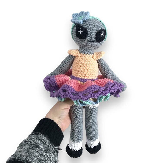A crocheted doll of a colorful alien doll with a gray body, wearing a multicolored skirt and a pale orange top, held in a person's hand.