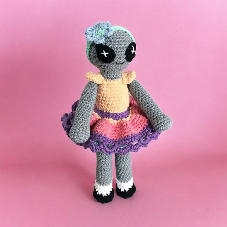A crocheted doll of a colorful alien doll with a gray body, wearing a multicolored skirt and a pale orange top, standing against a pink background.