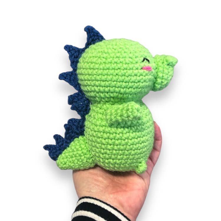 A small crochet baby dragon with a green body and blue spines, held in a person's hand.