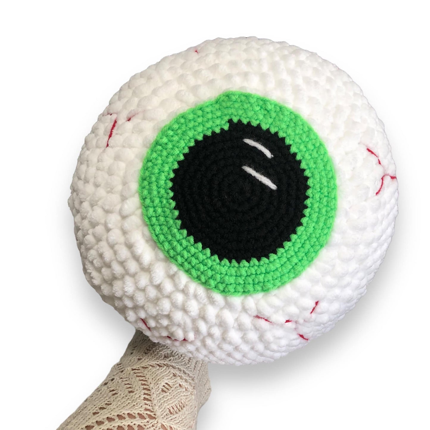 A large crocheted eyeball with a green iris being held up against a white background.