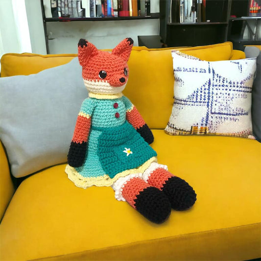 A crochet fox doll with a colorful dress and bloomers, sitting on a yellow couch with a pillow behind it.