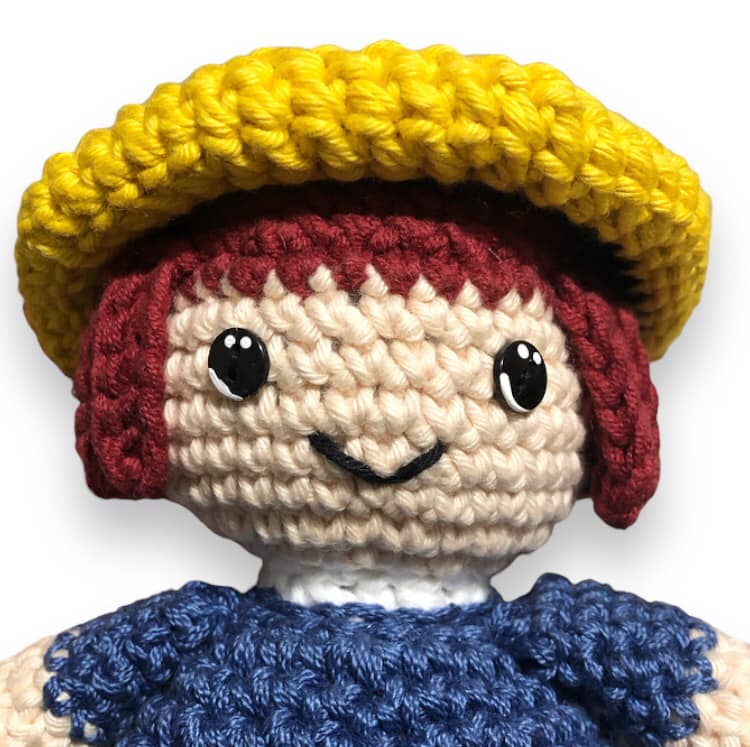 A close up of a crochet version of Madeline, from the children's book series, Madeline, against a white background.