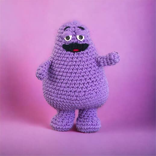 A crochet doll of Grimace standing against a purple background.
