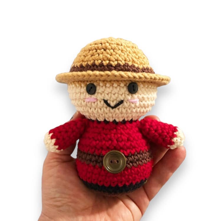 A crochet doll resembling a Canadian Mountie being held against a white background.