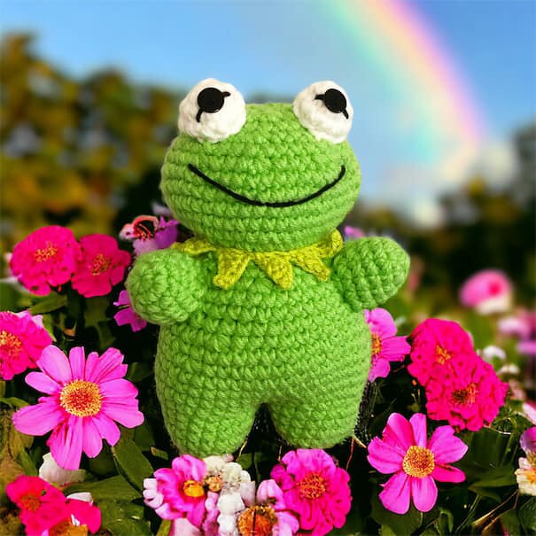 A crocheted doll resembling Kermit the Frog standing amongst some pink flowers.