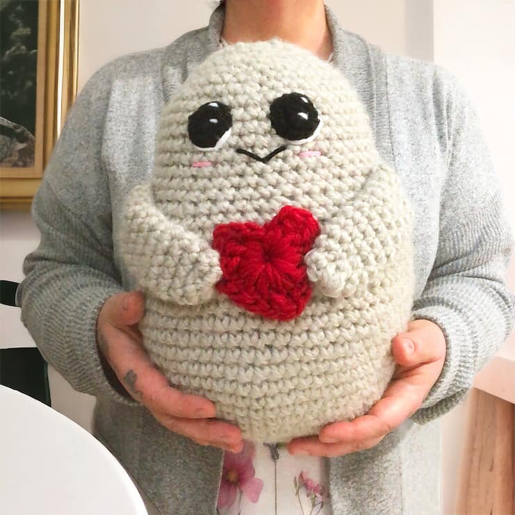 A crocheted potato with a red heart and large eyes being held up by a person.