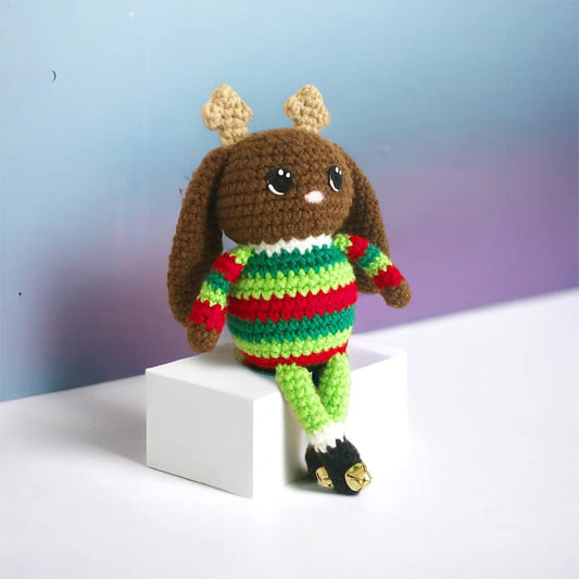 A crocheted jackalope doll with small antlers and a green and red striped sweater with little black boots that have bells on them sitting on a small white box.