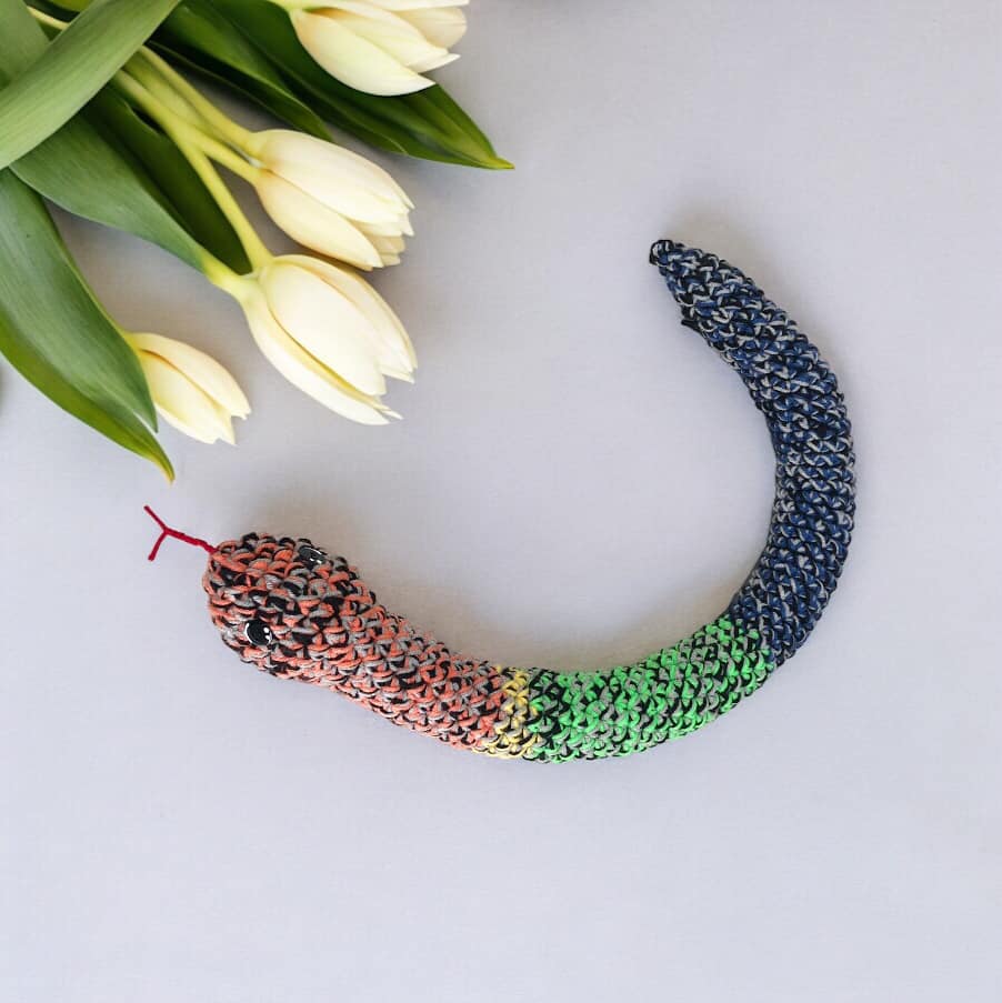 A crocheted rainbow rattle snake laid out next to white flowers.