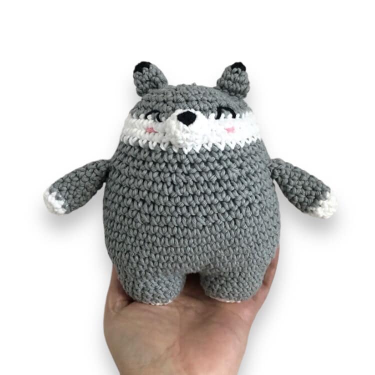 A crochet doll resembling a chubby wolf being held up against a white background.
