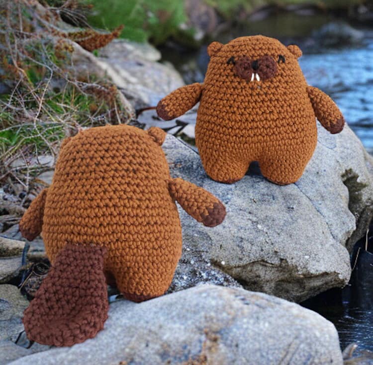 A crochet pattern for a beaver plush toy, showing two beavers made from brown yarn, sitting on a rock beside a stream.