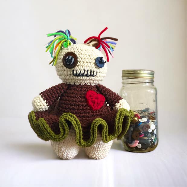 crocheted creepy doll standing next to a pair jar of buttons with a white background.