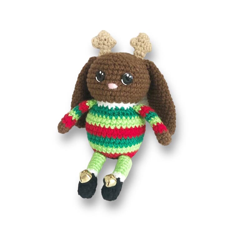 A crocheted jackalope doll with small antlers and a green and red striped sweater with little black boots that have bells on them sitting against a white backgound.