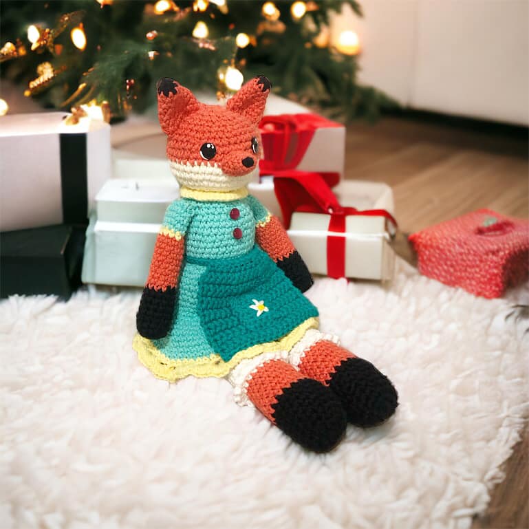 A crochet fox doll in a blue dress and bloomers sitting on a white carpet next to some presents under a Christmas Tree.