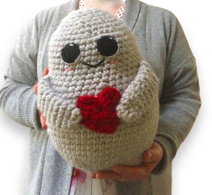 A crocheted potato with a red heart and large eyes being held up by a person.