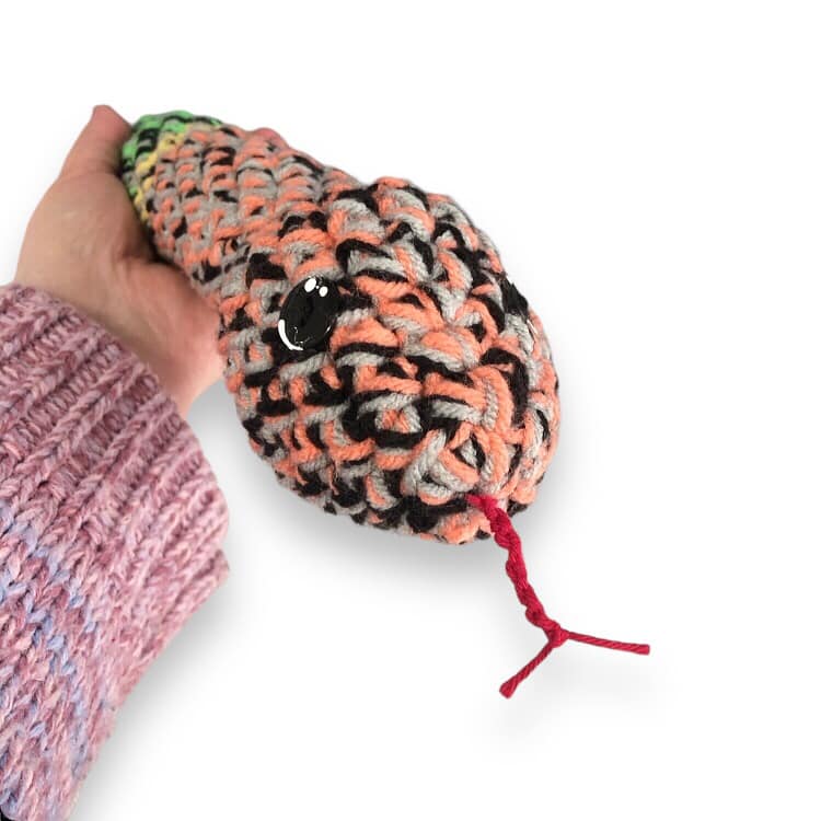 A close up of the rainbow crochet rattle snake, being held up by a hand against a white background.