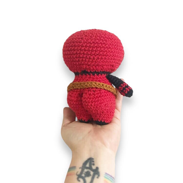 A behind view of the crochet Deadpool doll with bubble buttcheeks being held up against a white background.