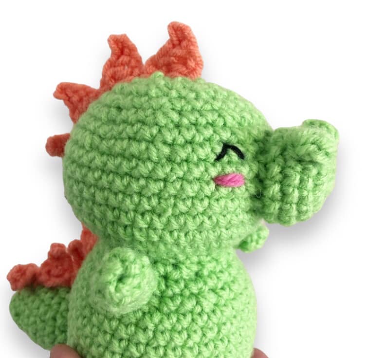 A small crochet baby dragon with a green body and orange spines, held in a person's hand.