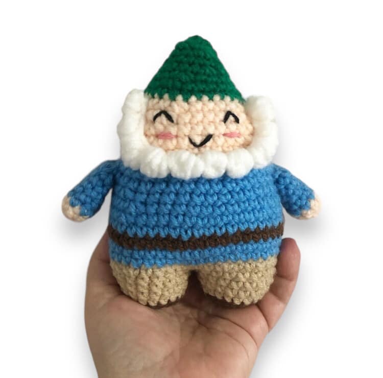 A crocheted doll of a smiling garden gnome with a green hat, blue jacket, beige pants, and a white beard.