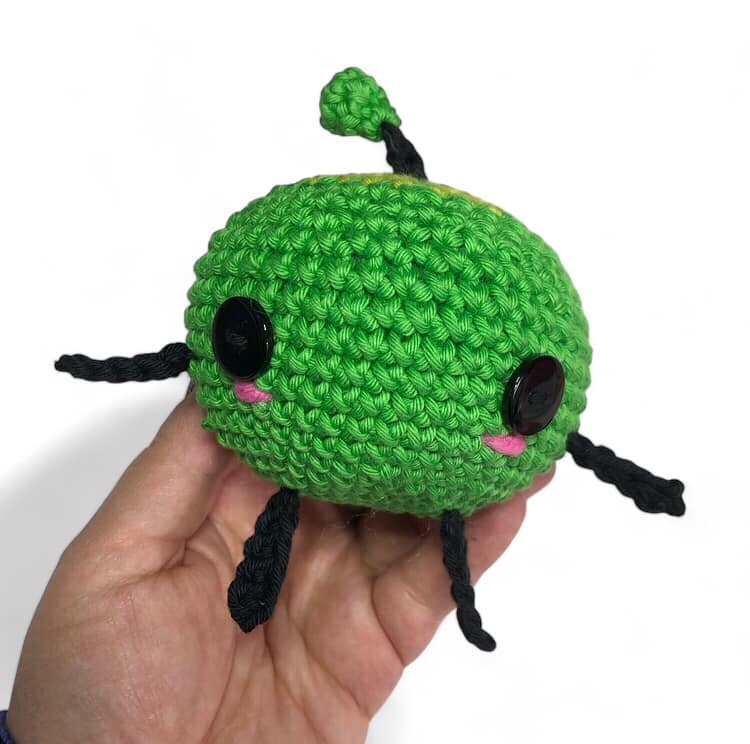 A green crochet Junimo being held up against a white background.