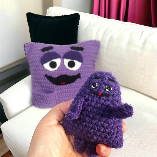 A crochet pattern for a purple Grimace cushion and a smaller crocheted doll of Grimace designed to fit into the pocket, displayed on a sofa.