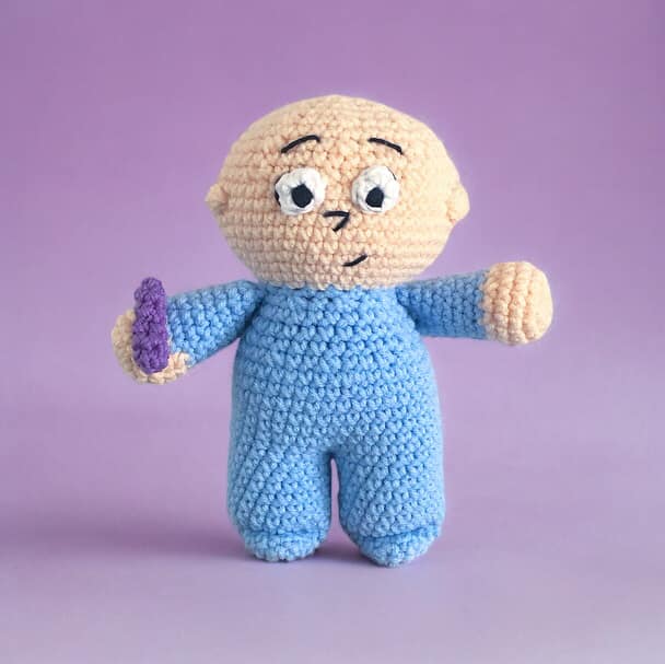 A crocheted doll resembling Harold from Harold and the Purple Crayon standing against a lavender background.
