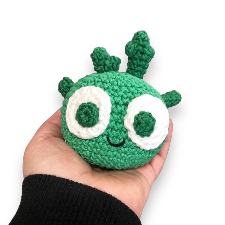 A crochet doll of a cartoon kohlrabi being held up against a white background.