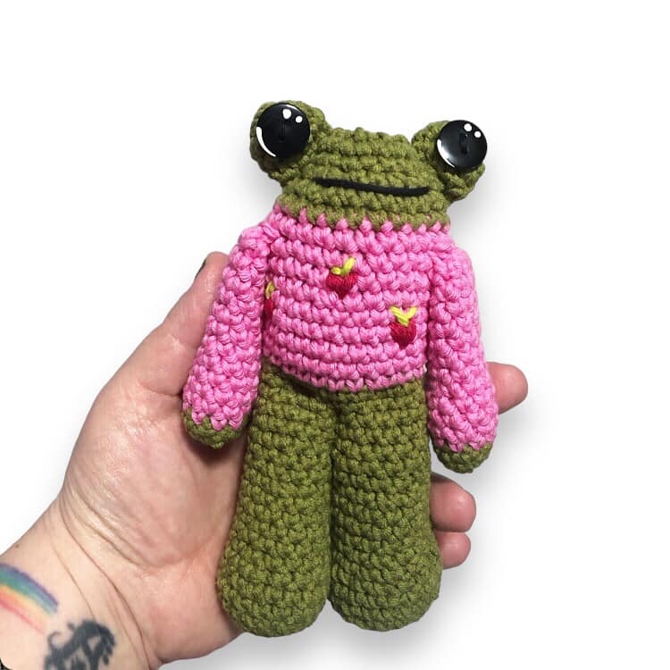 A crocheted frog doll in a pink sweater with strawberries being held up against a white background.
