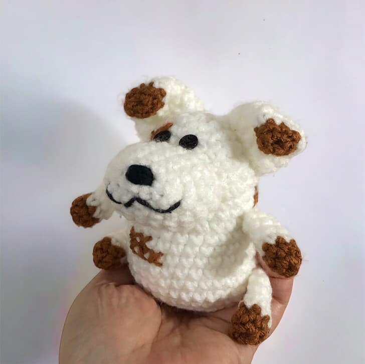 A crocheted dog doll resembling Russel from Nanalan being held up against a white background.