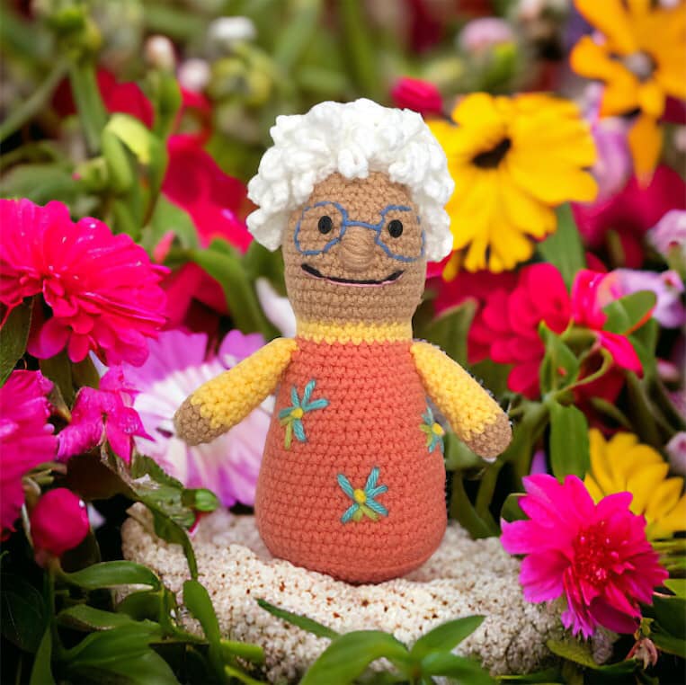 A crocheted doll resembling Nana from Nanalan standing against a field of flowers on a rock.