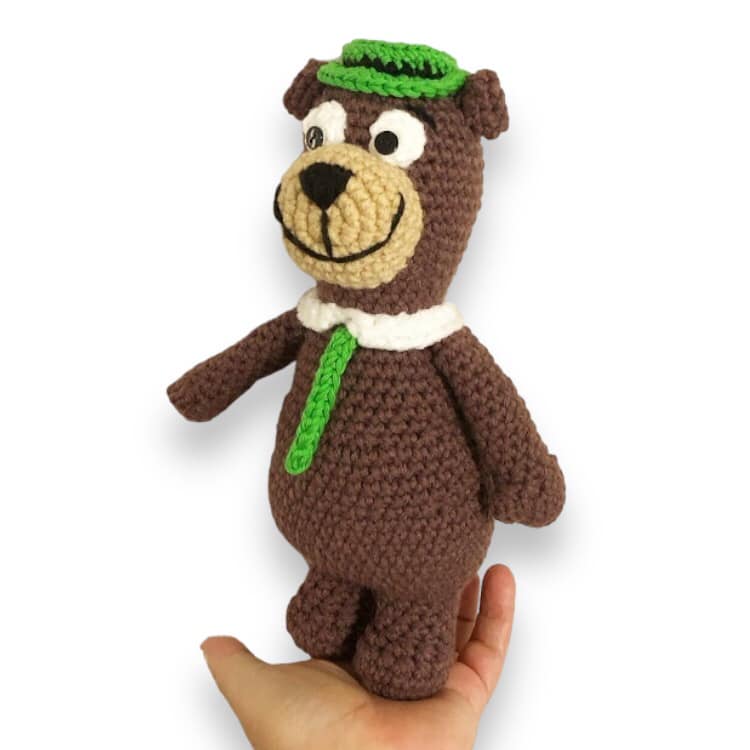 A crochet doll resembling Yogi Bear being held up against a white background.