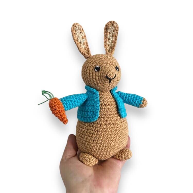 A crochet Peter Rabbit doll being held against a white background and holding a carrot with a blue coat