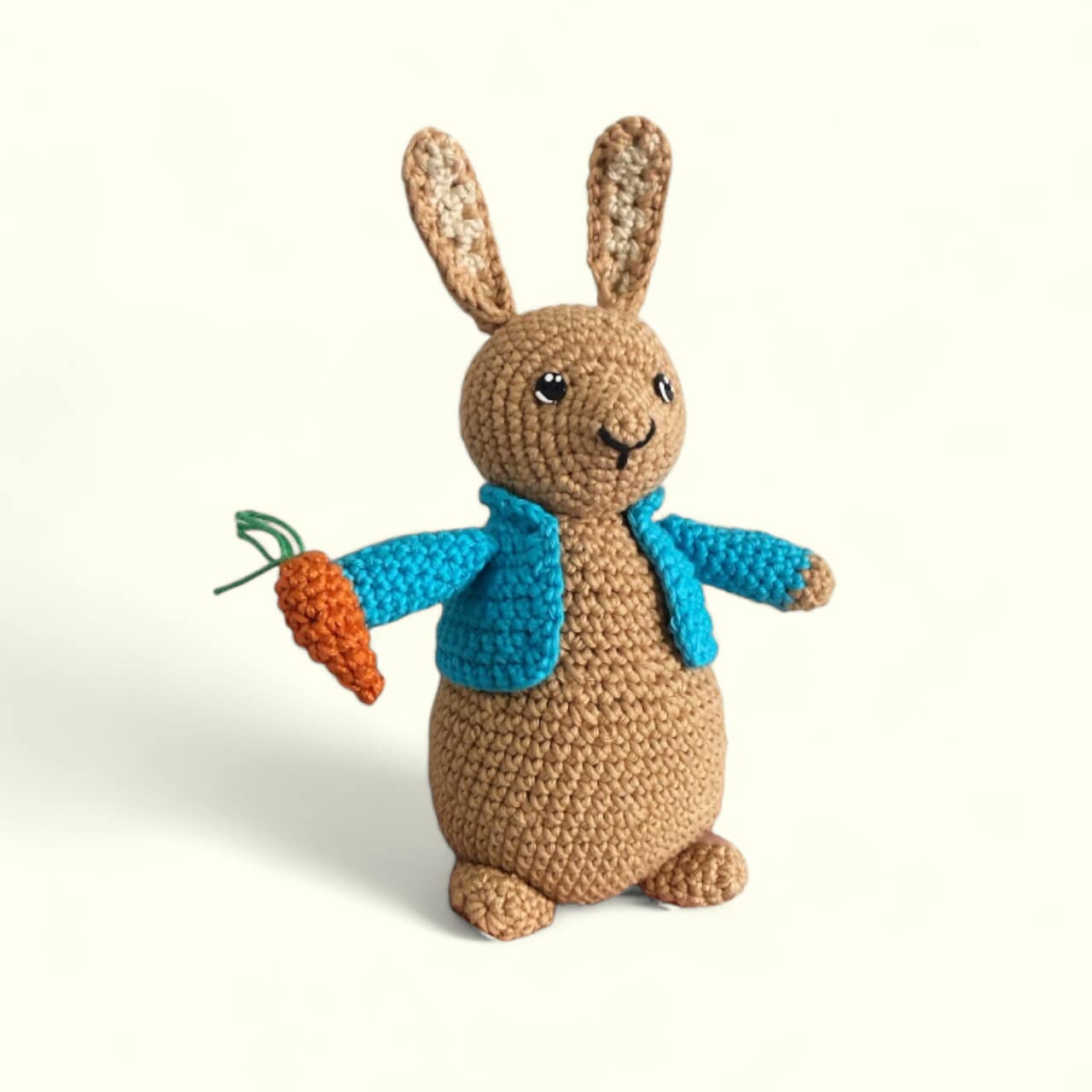 A crochet Peter Rabbit doll standing against a white background and holding a carrot with a blue coat