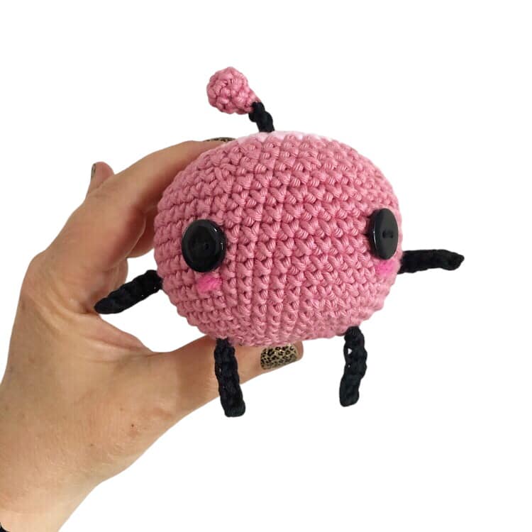 A pink crochet junimo being held up against a white background.