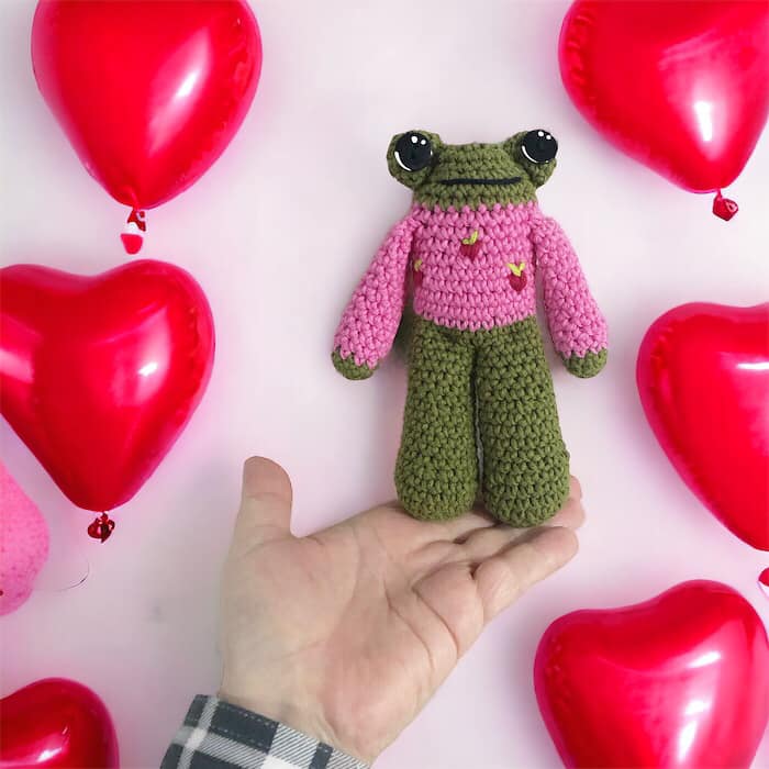 A crocheted plushie of a frog with a pink sweater, displayed alongside red heart-shaped balloons.