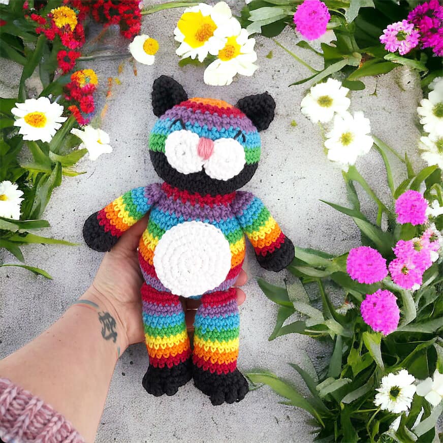 A crocheted rainbow cat being held against a background with flowers.