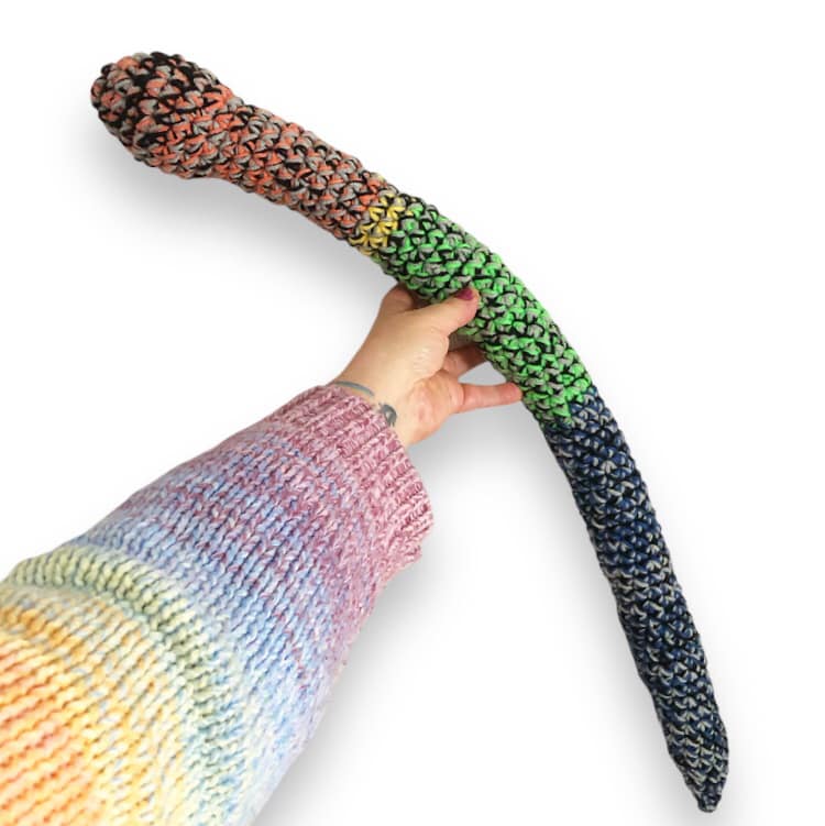 An image showing a colorful crochet snake with a rattle tail being held up by a hand against a white background.