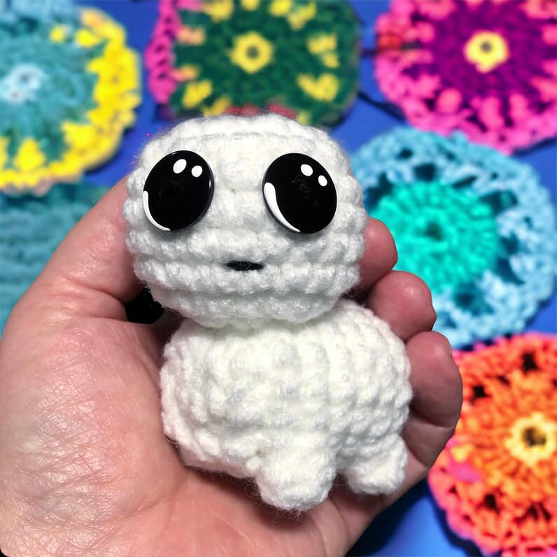 A small crochet creature with a white body and black eyes, held in a person's hand, with a variety of colorful crocheted items in the background.