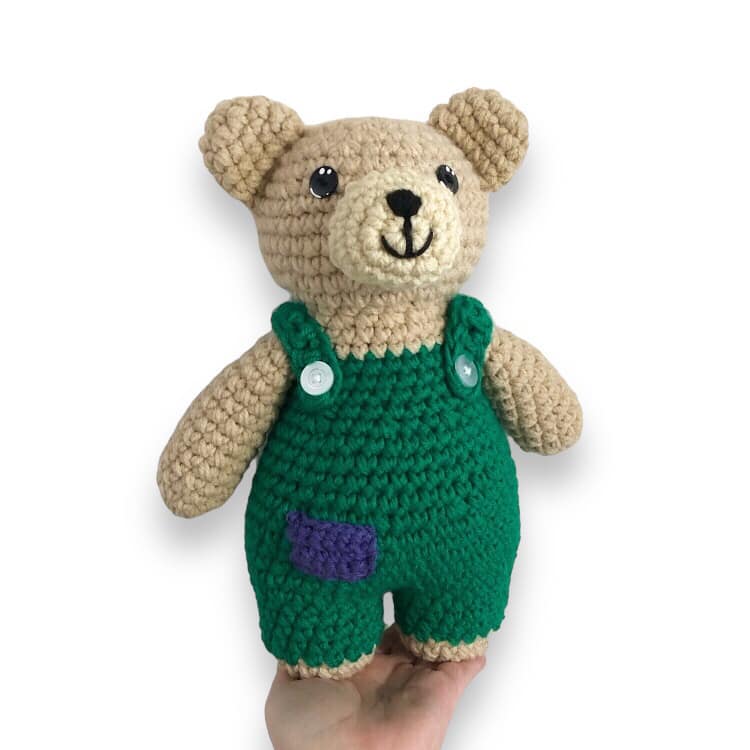 A crochet doll resembling Corduroy Bear being held against a white background.