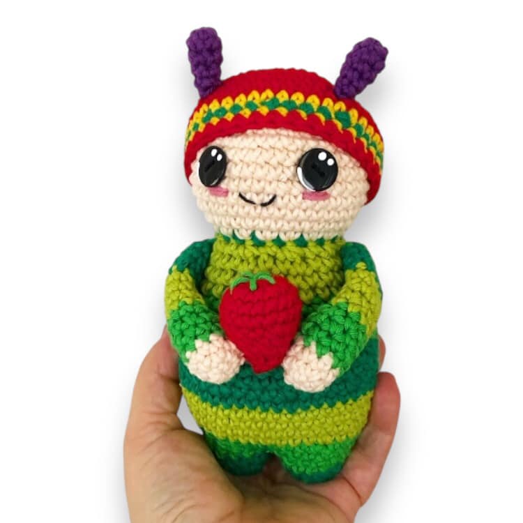 A crochet pattern for a toy resembling a small child dressed like caterpillar with a striped body, wearing a hat, and holding a strawberry. The image shows the finished toy and the pattern name 'Fat Lady Crochet'.