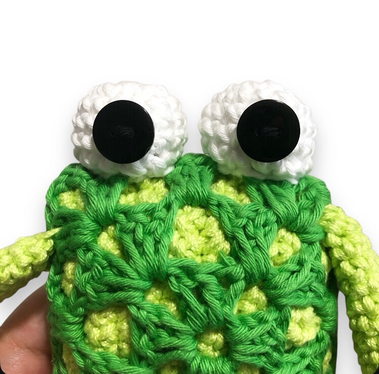 Crocheted frog toy with large eyes and no smile on a white background