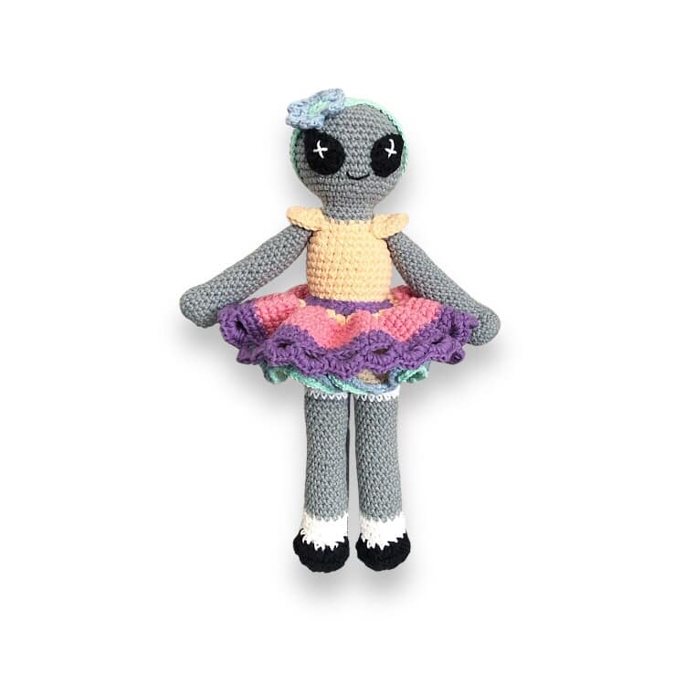 A crocheted doll of a colorful alien doll with a gray body, wearing a multicolored skirt and a pale orange top, standing against a white background. 