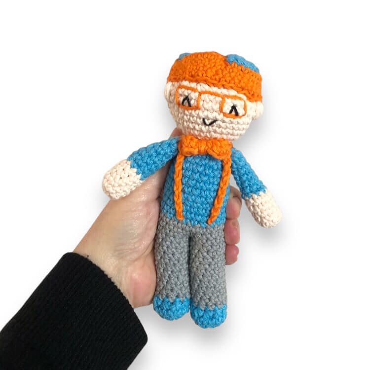 A crochet doll resembling Blippi with a orange hat, blue shirt, gray pants, and a bow tie.