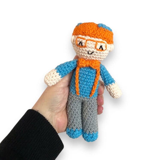 A crochet doll resembling Blippi with a orange hat, blue shirt, gray pants, and a bow tie.