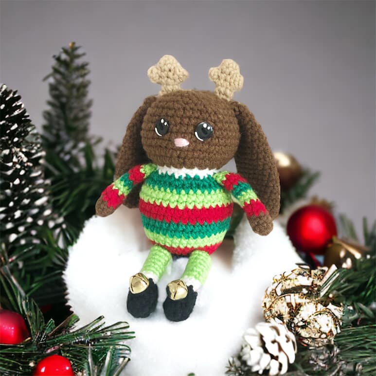 A crocheted jackalope doll with small antlers and a green and red striped sweater with little black boots that have bells on them sitting on a snowball surrounded by ornaments. 