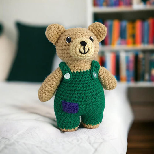 A crocheted doll resembling Corduroy Bear standing on a white blanket on a bed with a bookshelf in the background. 