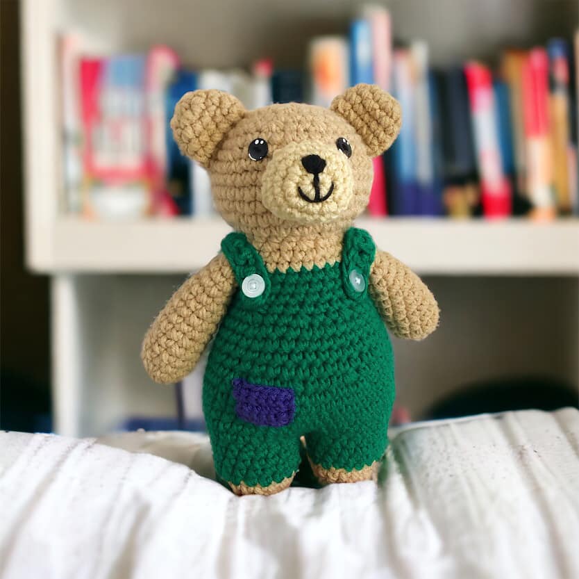 A crocheted doll resembling Corduroy Bear standing on a white blanket with books in the background. 