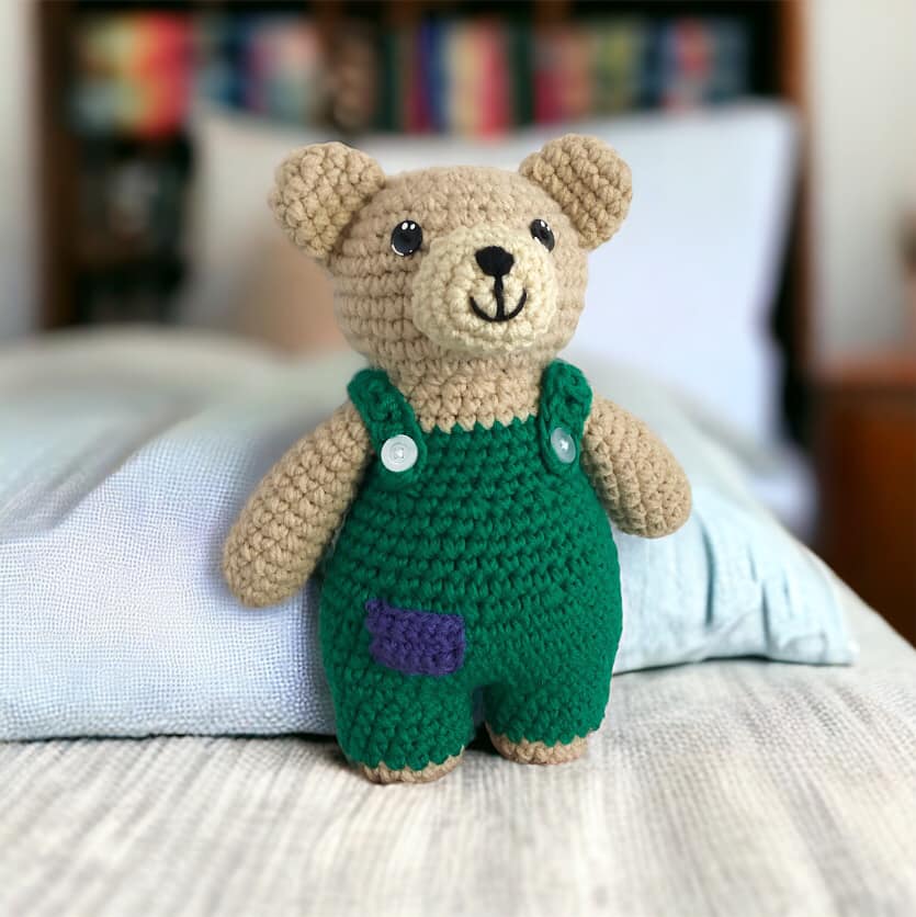 A crochet doll resembling Corduroy Bear standing on a bed. 