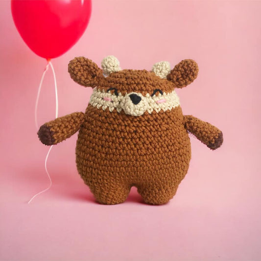 A crochet doll resembling a rotund deer standing against a pastel pink background with a hot pink balloon. 