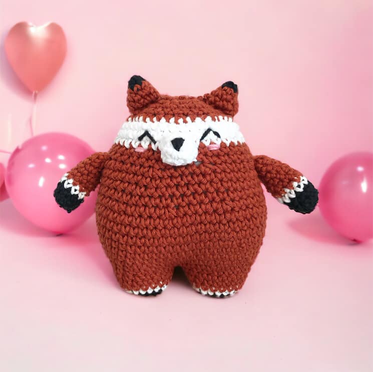 A crocheted doll resembling a chubby fox standing against a pink background with pink balloons. 