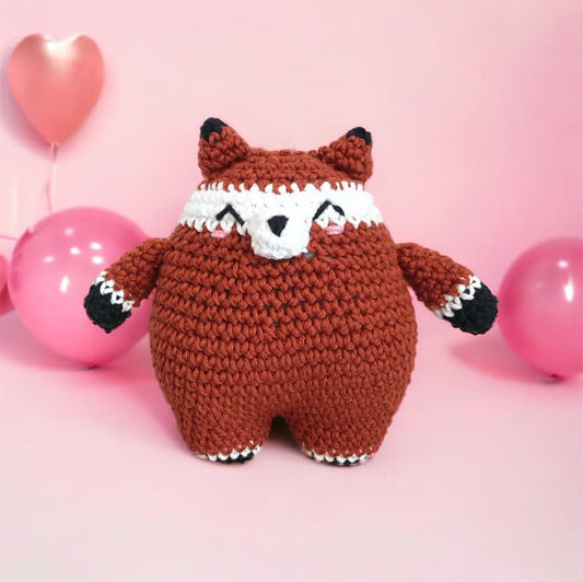 A crocheted doll resembling a chubby fox standing against a pink background with pink balloons. 